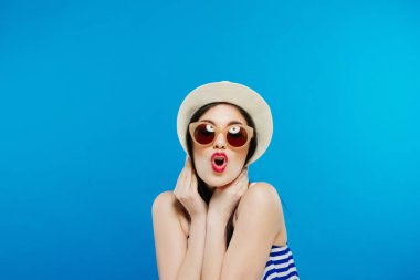 Şapka ve güneş gözlüklü neşeli güzel kız. Yaygın olarak gülümseyen, seyir ve bir kenara göstererek. Yaz kıyafeti. Belden yukarı, stüdyo, kapalı