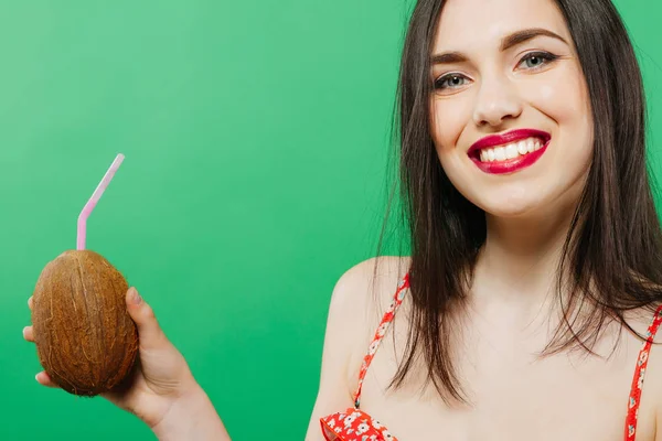 Retrato de una joven sonriente bebiendo cóctel tropical en coco y mirando a la cámara sobre fondo verde . — Foto de Stock