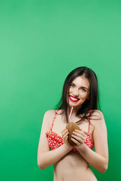 Increíble morena sonriente en traje de baño brillante posando con cóctel en el estudio sobre fondo verde . — Foto de Stock