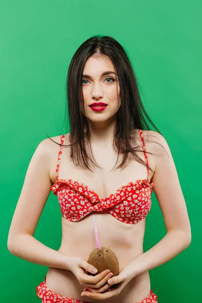 Morena con el pelo largo en traje de baño rojo sosteniendo coco en sus manos en el estudio sobre fondo verde . — Foto de Stock