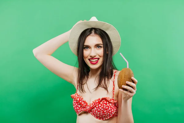 Retrato de la morena con sombrero de vaquero y pelo largo en traje de baño rojo sosteniendo coctel de coco en sus manos en el estudio sobre fondo verde . — Foto de Stock