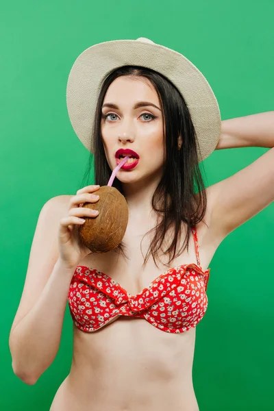 Increíble morena en traje de baño brillante y sombrero de vaquero posando con cóctel en el estudio sobre fondo verde . — Foto de Stock