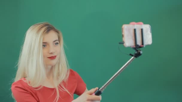 Geweldige Blonde nemen foto via Selfie Stick op groene achtergrond. Schattig meisje in rood Shirt fotograferen zelf door Smartphone in Studio. — Stockvideo