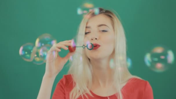 Portret van grappige mooie meisje een heleboel bellen blazen op groene achtergrond. Studio Shot van Blonde met lange haren en sensuele lippen dragen rode Shirt. — Stockvideo