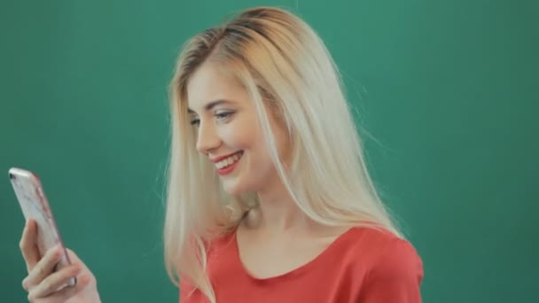 Mulher loira bonito está olhando em seu smartphone e sorrindo em fundo verde no estúdio. Retrato de close-up de muito jovem usando celular . — Vídeo de Stock