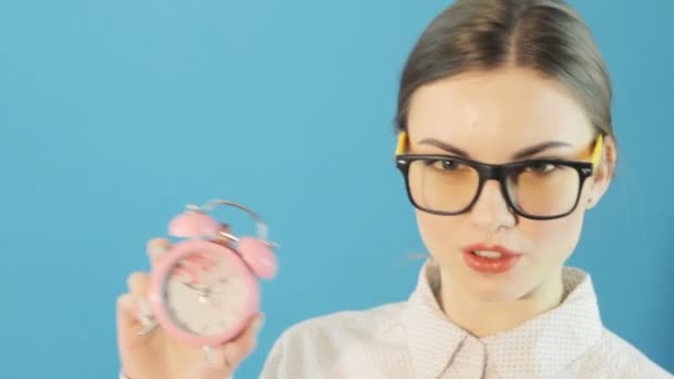 Hermosa morena con cola de caballo en gafas y camisa sosteniendo reloj rosa en las manos sobre fondo azul brillante en el estudio. Concepto de tiempo . — Vídeo de stock