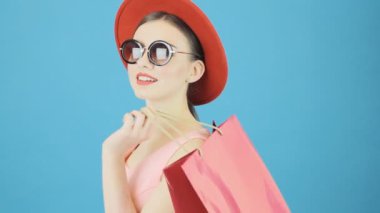 Şirin esmer kırmızı şapka ve güneş gözlüğü Studio mavi arka plan üzerinde pembe alışveriş çantası elinde. Mevsimlik satış konsepti.