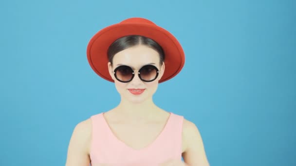 Porträt einer brünetten Shopperin mit rotem Hut und Sonnenbrille, die im Studio eine rosa Einkaufstasche auf blauem Hintergrund hält. Saisonales Verkaufskonzept. — Stockvideo