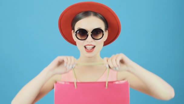 Primer plano Retrato de una mujer morena con sombrero rojo y gafas de sol frescas sosteniendo una bolsa de compras rosa sobre fondo azul en el estudio. Concepto de venta estacional . — Vídeo de stock