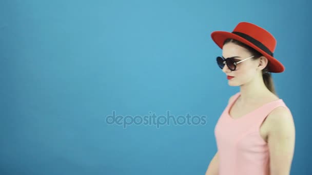 Портрет симпатичной брюнетки на голубом фоне в студии. Молодая женщина в красной шляпе и солнцезащитных очках смотрит в камеру . — стоковое видео