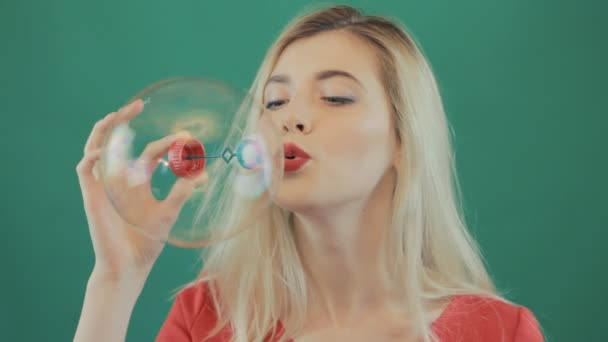 Portrait of Blonde with Long Hair and Sensual Lips Wearing Red Shirt Playing in Studio. Playful Young Girl Blowing Big Bubbles on Green Background. — Stock Video