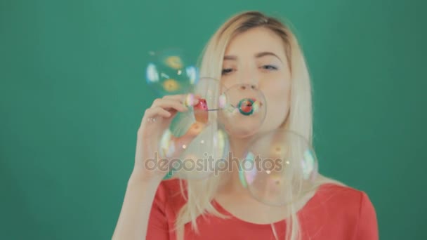 Lekfull kvinna blåser bubblor på kameran på grön bakgrund. Studio skott av blond med långt hår och sensuella läppar iklädd röd skjorta. — Stockvideo