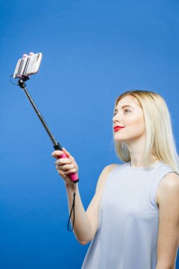 Neşeli sarışın mavi arka plan üzerinde Selfie Stick kullanarak fotoğraf çekiyorum. Sevimli kız elbiseli kendini Smartphone Studio tarafından fotoğraf çekimi.