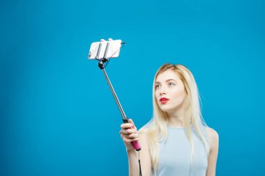 Şehvetli dudakları ile şirin sarışın kendini fotoğraf çekme. Gülümseyen kız mavi arka plan üzerinde bir fotoğraf çekmek için Selfie Stick kullanarak.