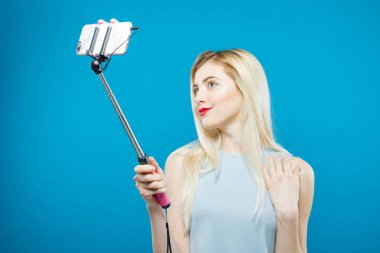 Şehvetli dudakları ile şirin sarışın kendini çekim portresi. Gülümseyen kız mavi arka plan üzerinde bir fotoğraf çekmek için Selfie Stick kullanarak.