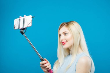 Harika sarışın şehvetli dudakları ve vahşi gülümseme kendini fotoğraf çekimi ile. Gülümseyen kız mavi arka plan üzerinde bir fotoğraf çekmek için Selfie Stick kullanarak.