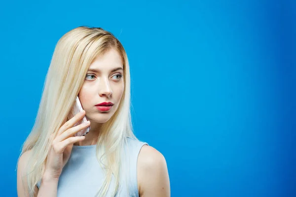 Lindo vestido rubio usando está llamando a alguien usando su teléfono inteligente sobre fondo azul. Chica atractiva con móvil en el estudio . — Foto de Stock