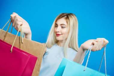 Şehvetli sarışın renkli çanta alışveriş çok. Milano saç ve çekici gülümseme Studio mavi arka plan ile mutlu kız.