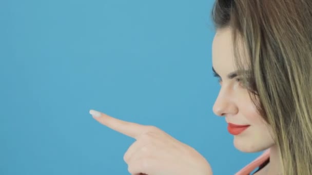 Menina incrível com cabelo escuro longo está posando no fundo azul e mostrando por dedo em algo . — Vídeo de Stock
