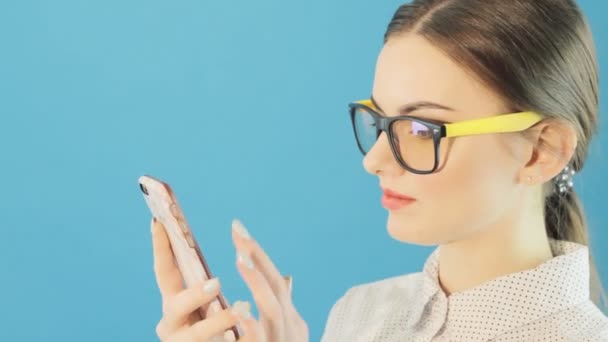 Atrakcyjne Hipster Girl ubrany modne okulary i jej smartfona do znaleźć informacje w Internecie. Brunetka z urządzenia mobilnego korzysta ekran dotykowy na niebieskim tle w Studio. — Wideo stockowe