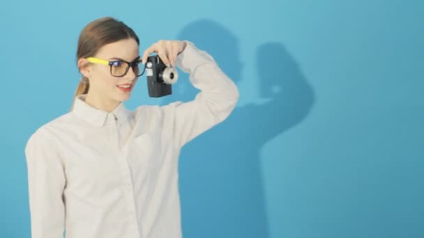 Retrato de la hermosa morena con gafas y camisa está utilizando la cámara retro sobre fondo azul en el estudio. Mujer joven tomando fotos . — Vídeos de Stock