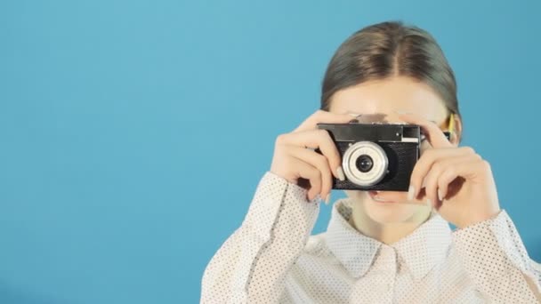 Primer plano retrato de la linda morena con gafas y camisa está utilizando la cámara retro sobre fondo azul en el estudio . — Vídeo de stock