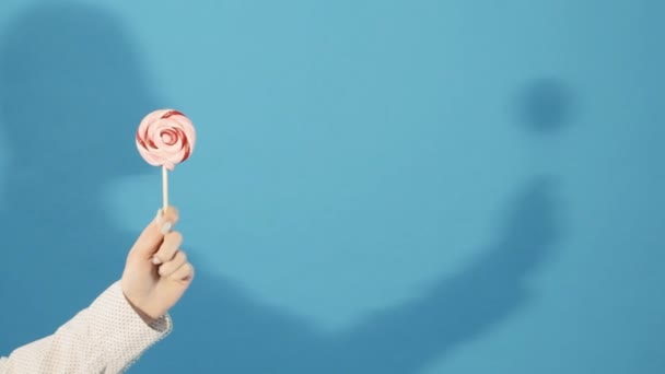 Female Hand Holding Colorful Lollipop on Blue Background with Girl Shadow in Studio. — Stock Video