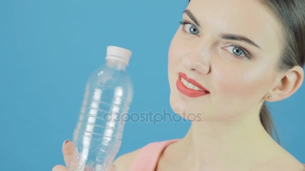 Sonriente linda morena con botella de agua mineral sobre fondo azul en el estudio . — Vídeo de stock