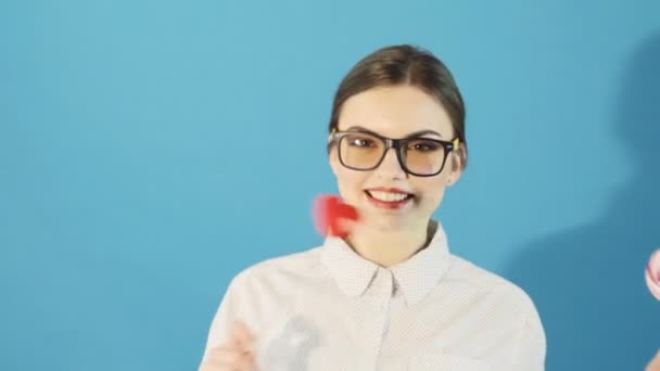 Porträt eines süßen, gut aussehenden Mädchens mit Pferdeschwanz und Brille, das Herz und Lutscher in den Händen hält und im Studio auf blauem Hintergrund posiert. lustige Brünette mit roten Lippen im Hemd. — Stockvideo