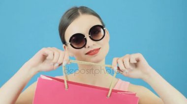 Pembe alışveriş çantası Studio mavi arka plan üzerinde tutarak serin güneş gözlüklü esmer kadın. Mevsimlik satış konsepti.