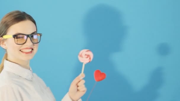 Porträt eines lustigen Mädchens mit Pferdeschwanz und Brille, das Herz und Lutscher in den Händen hält und im Studio auf blauem Hintergrund posiert. niedliche Brünette mit roten Lippen im Hemd. — Stockvideo