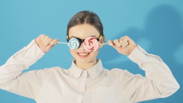 Retrato de una joven con cola de caballo y gafas sosteniendo dos piruletas en las manos posando en el estudio sobre fondo azul. Linda morena con labios rojos en camisa . — Vídeos de Stock