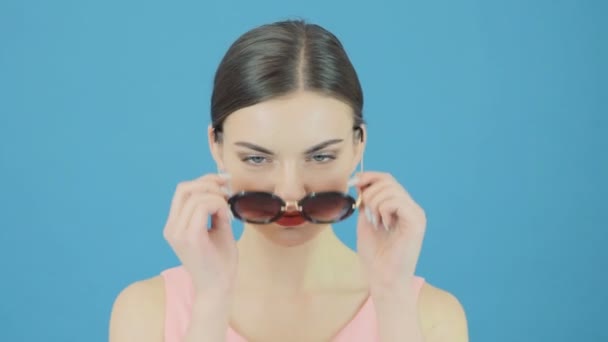 Retrato de moda de la modelo morena con gafas de sol frescas y vestido rosa sobre fondo azul. Chica perfecta está posando en el estudio . — Vídeo de stock