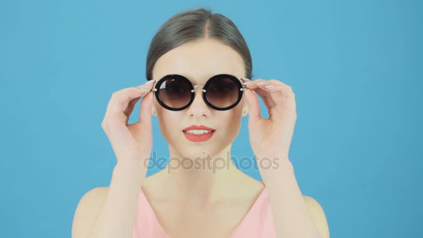 Retrato de belleza de la morena modelo con gafas de sol de moda y vestido rosa sobre fondo azul. Chica perfecta está posando en el estudio . — Vídeo de stock