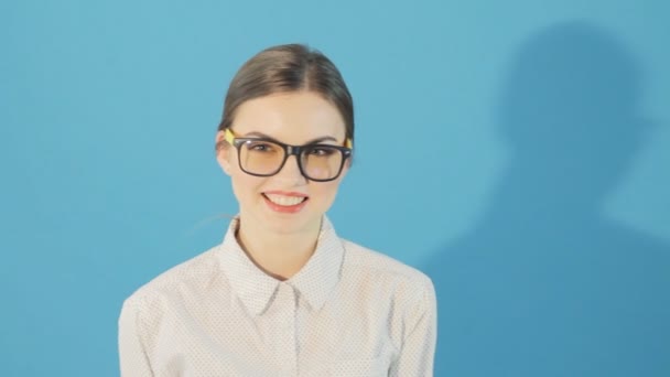 Jeune fille avec queue de cheval et lunettes montrant deux sucettes l'une après l'autre en studio sur fond bleu. Brune mignonne avec lèvres rouges en chemise . — Video