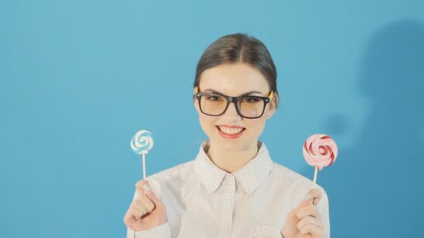 Morena usando gafas está sosteniendo dos piruletas en las manos y posando en el estudio sobre fondo azul . — Vídeo de stock