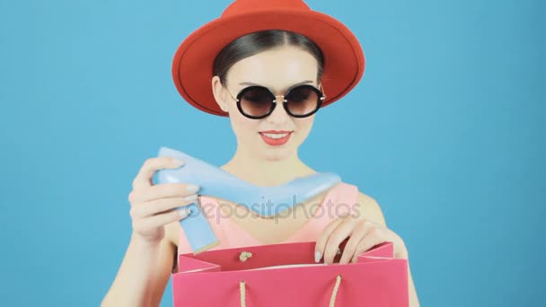 Vackra brunett kvinnlig Shopper bär röd hatt och solglasögon är Holding shoppingkassar och blå sko. Säsongsbetonad försäljning koncept. — Stockvideo