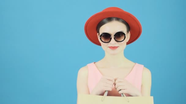 Schattige Brunette in Red Hat en coole zonnebril is boodschappentas Holding in handen. Seizoensgebonden verkoop Concept. Geïsoleerd op blauwe achtergrond in de Studio. — Stockvideo