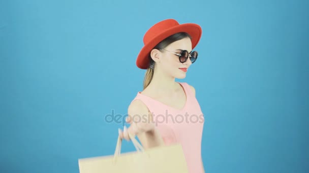 Fantastisk kvinna med röd hatt och solglasögon på blå bakgrund i Studio. Glad brunett håller en hel del Shopping färgglada väskor. Säsongsbetonad försäljning koncept. — Stockvideo