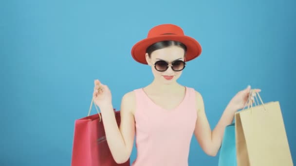Harika esmer kırmızı şapka ve güneş gözlüğü Studio mavi arka plan ile. Renkli çanta alışveriş bir sürü tutan mutlu kadın. Mevsimlik satış konsepti. — Stok video
