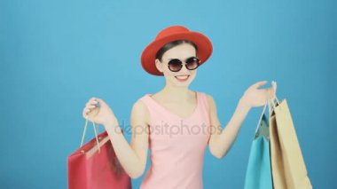 Portre sevimli esmer kırmızı şapka ve güneş gözlüğü Studio mavi arka plan ile. Renkli çanta alışveriş bir sürü tutan mutlu kadın. Mevsimlik satış konsepti.