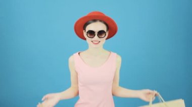 Komik kadın Shopper giydiği kırmızı şapka ve güneş gözlüğü Holding alışveriş torbaları Studio mavi arka plan üzerinde var. Mevsimlik satış konsepti.