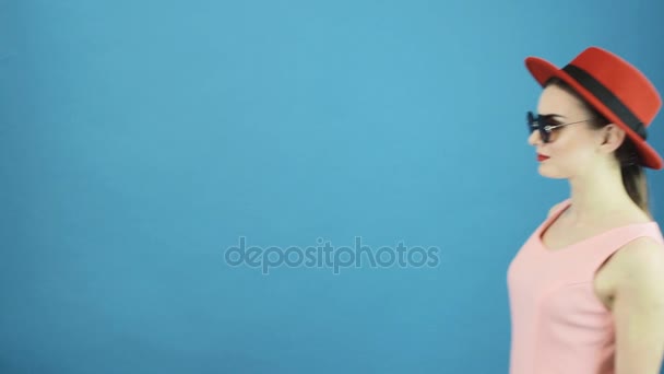 Retrato de la hermosa chica sobre fondo azul en el estudio. Increíble mujer joven con sombrero rojo y gafas de sol mirando a la cámara y alejándose . — Vídeos de Stock