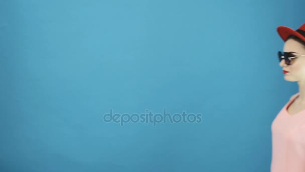 Amazing Young Woman Wearing Red Hat and Sunglasses Looking at the Camera and Walking Away. Portrait de belle fille sur fond bleu en studio .