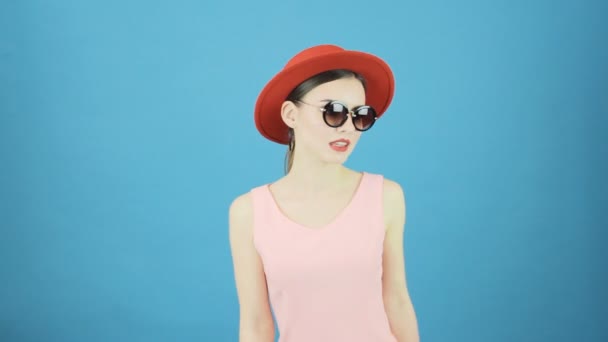 Schattige Brunette op blauwe achtergrond in de Studio. Portret van de jonge vrouw die het dragen van de rode hoed en zonnebril kijken naar de Camera. — Stockvideo