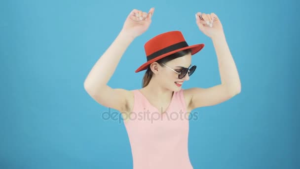 Verspieltes brünettes Mädchen mit rotem Hut und Sonnenbrille tanzt auf blauem Hintergrund im Studio. — Stockvideo