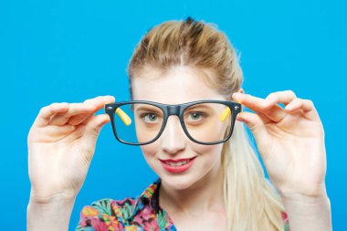 Closeup eğlence gülümseyen mavi arka plan üzerinde renkli gömlek ve moda gözlük takıyor at kuyruğu ile sarışın kadın portresi. Studio Posing şehvetli sevimli kızdır.