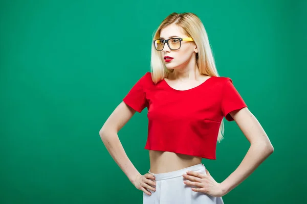 Retrato de chica sorprendida usando top corto rojo y gafas graduadas. Sensual guapa rubia con el pelo largo está posando sobre fondo verde en el estudio . — Foto de Stock