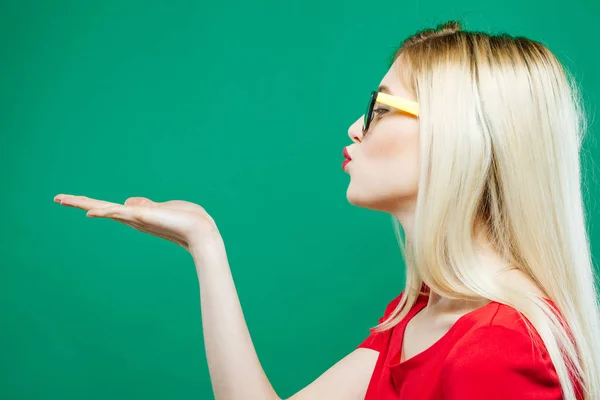 Vista laterale della giovane donna con lunghi capelli biondi, occhiali da vista e top rosso che tiene lo spazio vuoto sulla mano e si bacia su sfondo verde in studio . — Foto Stock