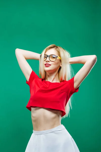 Девушка-блондинка с длинным каблуком и очками в белых юбках и красном топе в студии на зеленом фоне . — стоковое фото